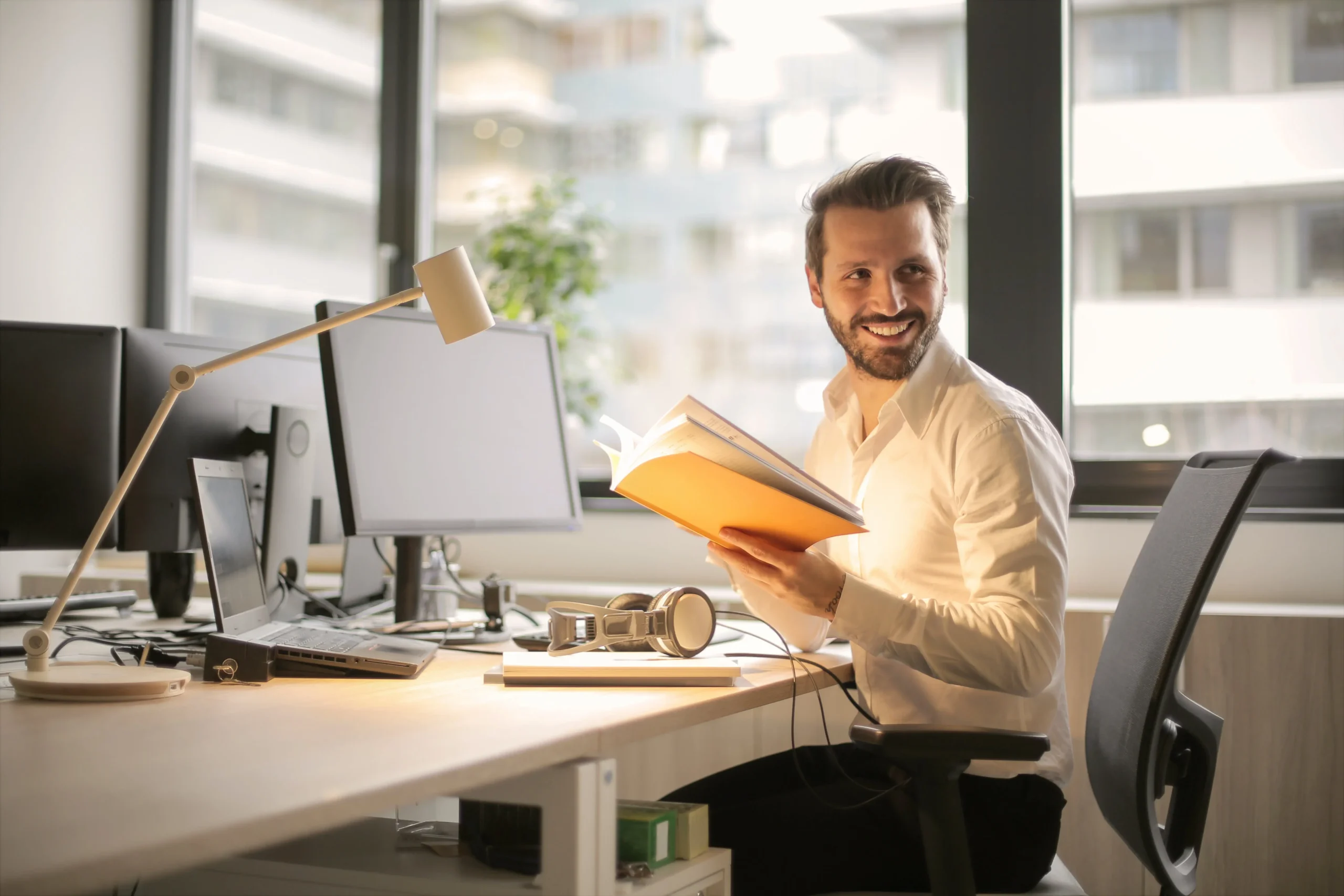 A smiling man holding a book while sitting at a desk