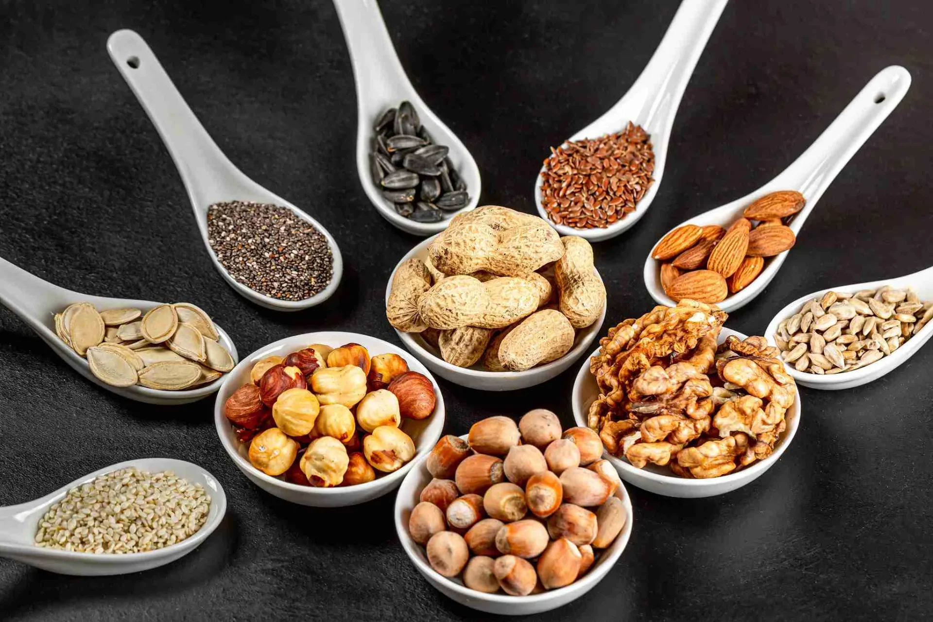 Assortment of nuts and seeds in white bowls and spoons on a black background